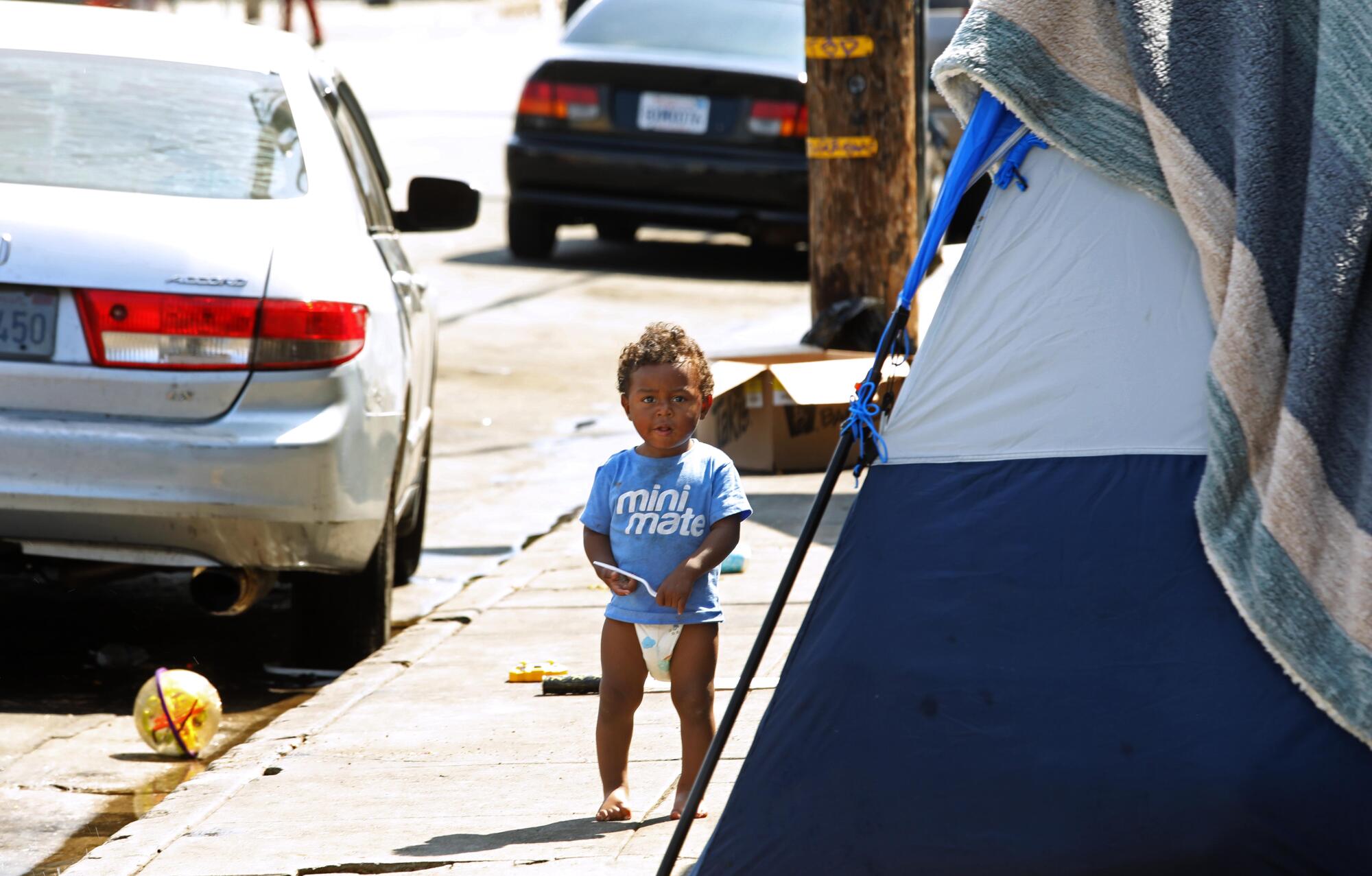 A migrant child in diapers stands barefoot in front of his family's tent 