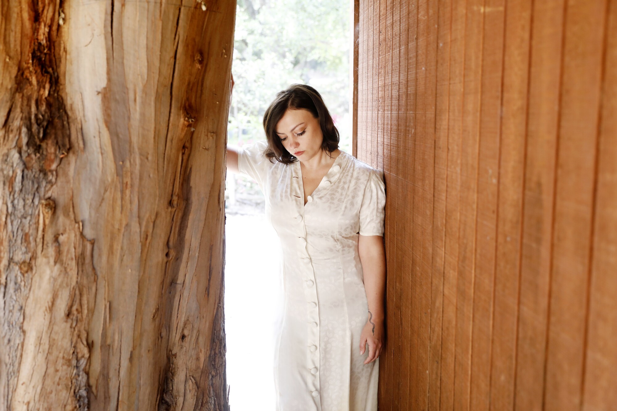 A woman in a white dress leans against a wooden wall and looks down, sullen