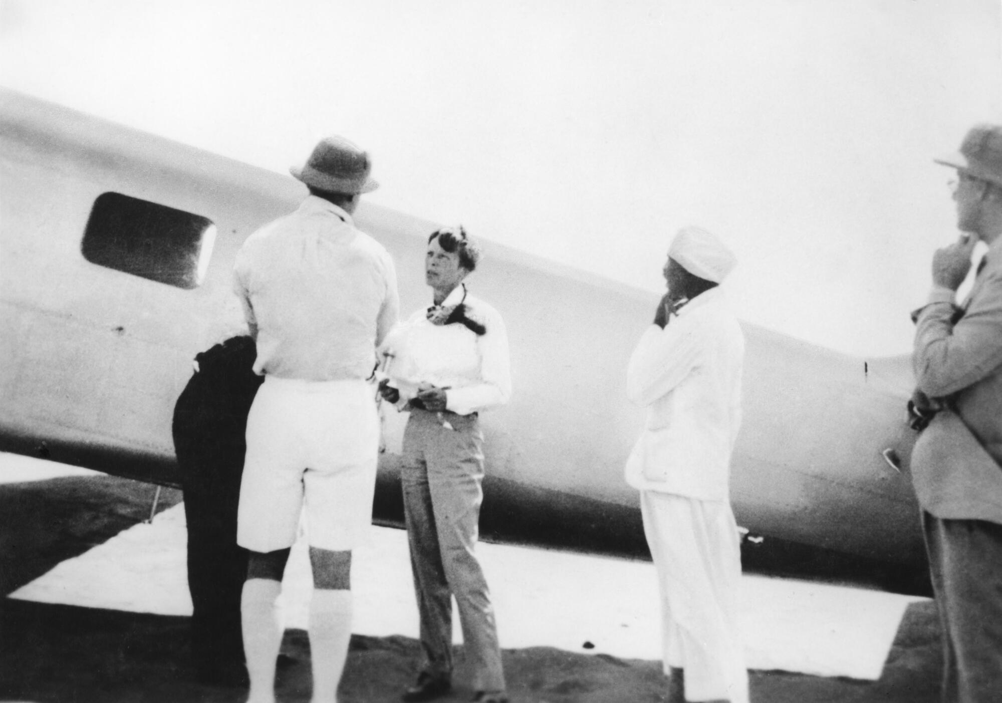 Amelia Earhart stands near an airplane fuselage with men in pith helmets.