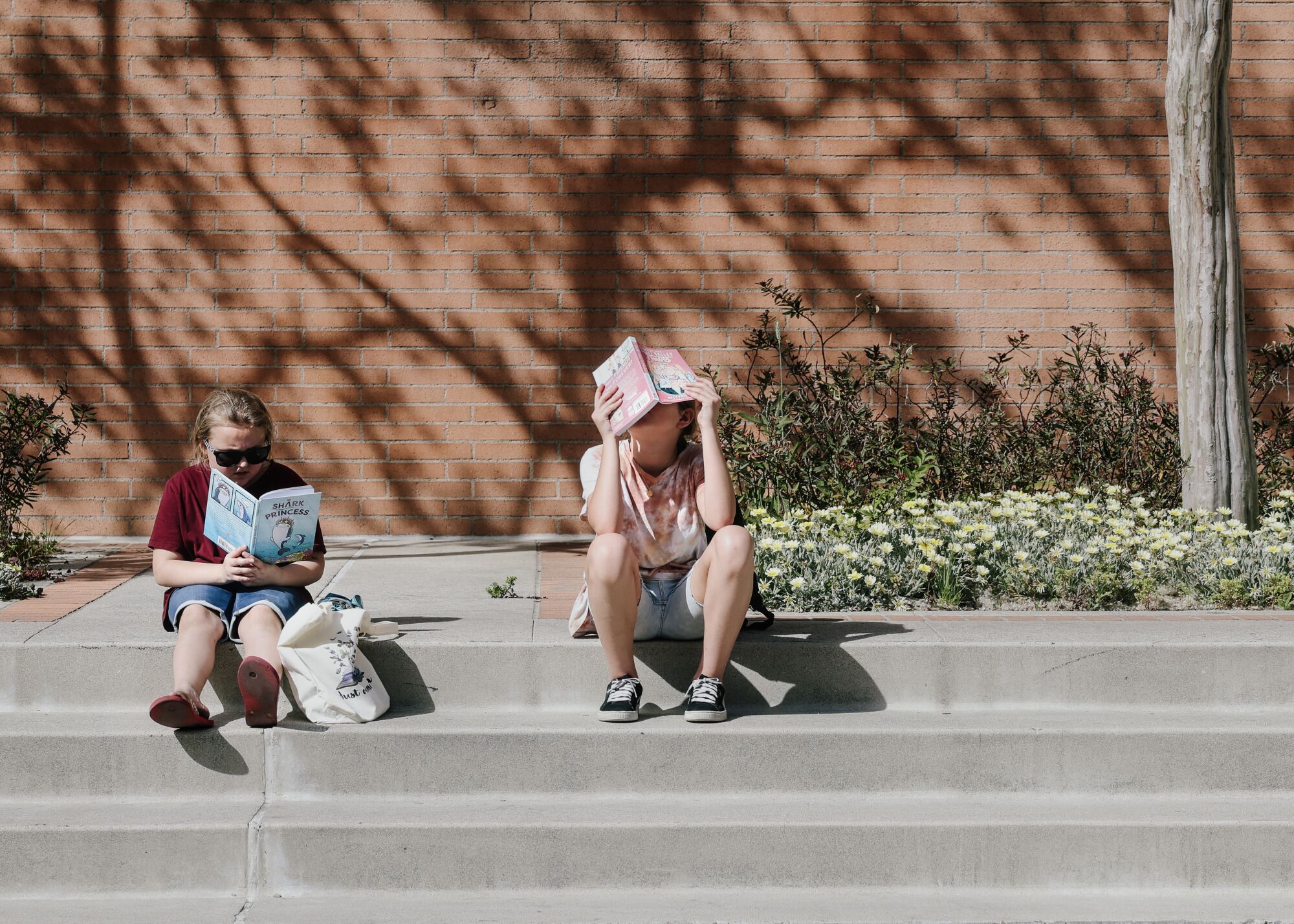 Açık havada beton basamaklarda oturan iki çocuk kitap okuyor.