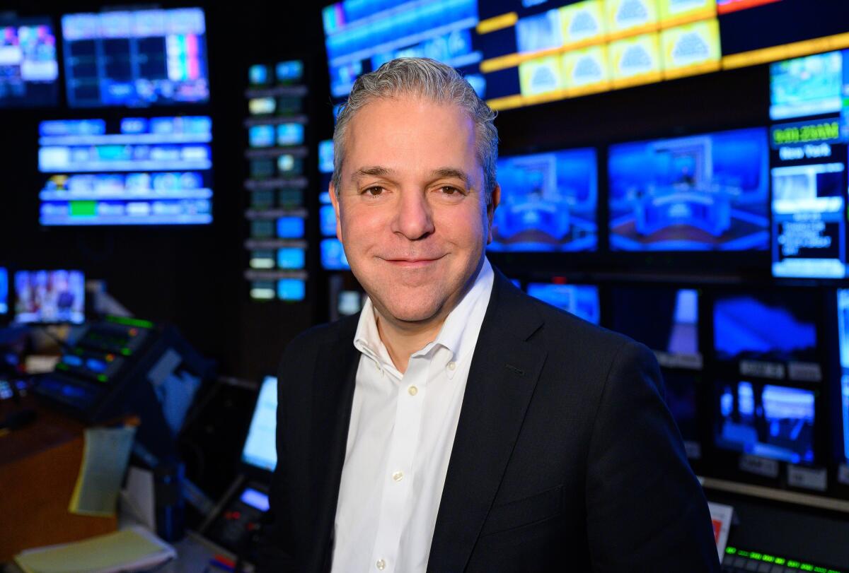 A man in a dark suit jacket and open-neck shirt with a bank of TV monitors behind him.