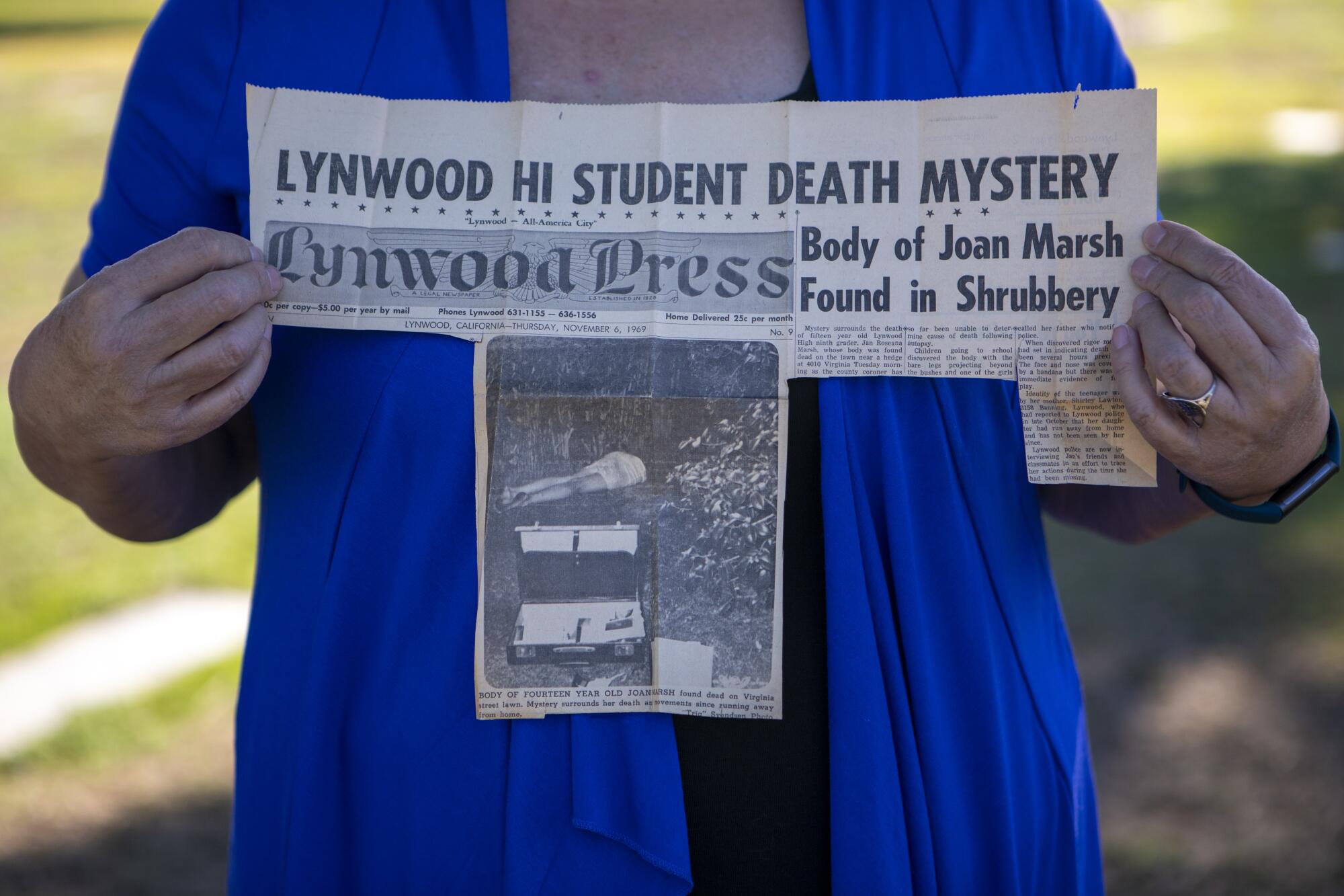 A woman holds a newspaper clipping that reads, "LYNWOOD HI STUDENT DEATH MYSTERY"
