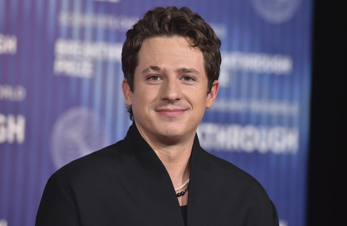 Charlie Puth standing with a pleasant face in front of a blue background while wearing a black jacket and necklaces.
