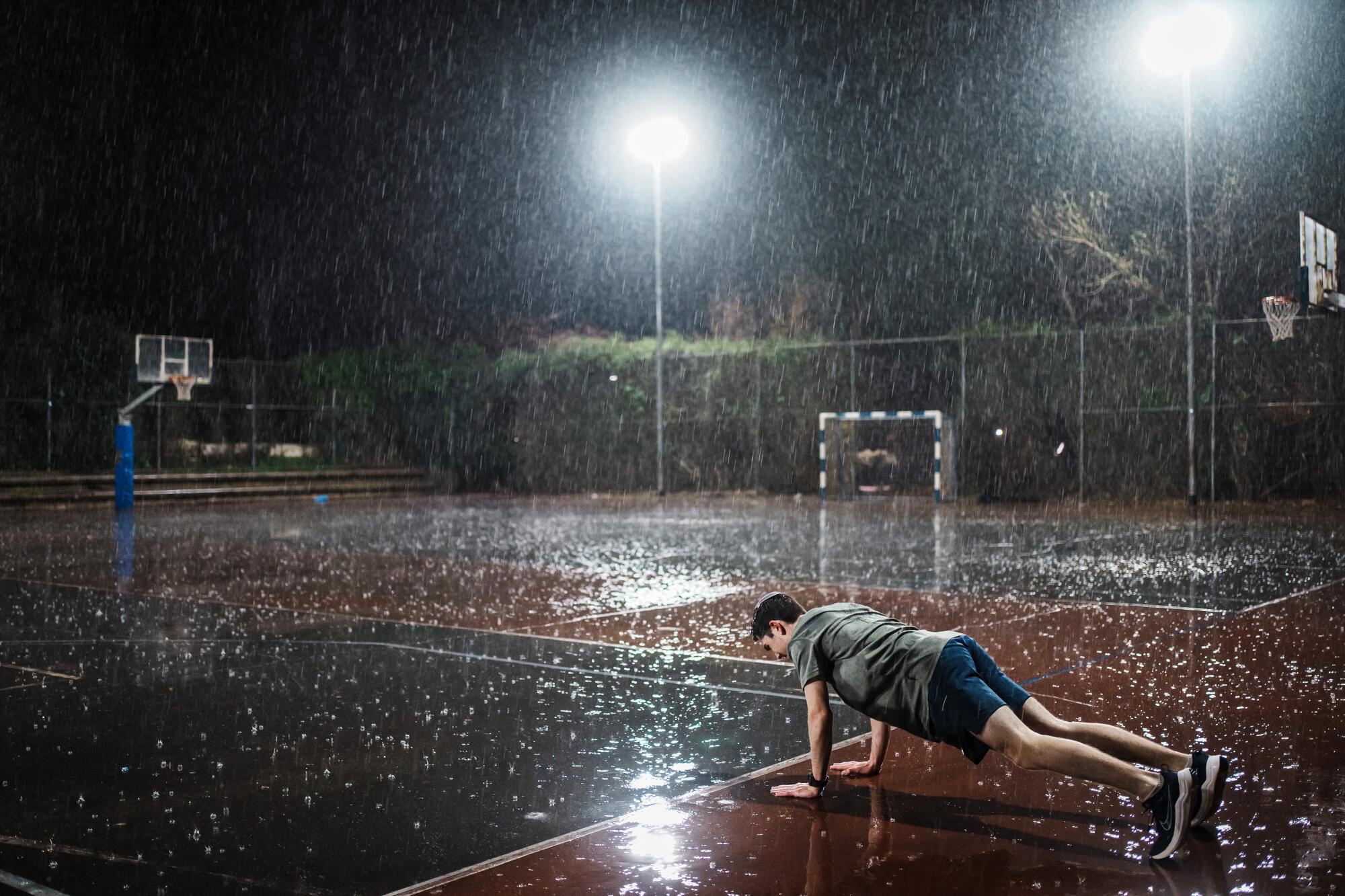 A young man wearing shorts does push-ups in the dark and rain on an athletic field. 