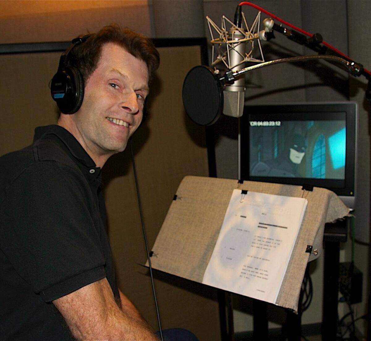 A man wearing headphones sits in front of a script and monitor in a voiceover booth