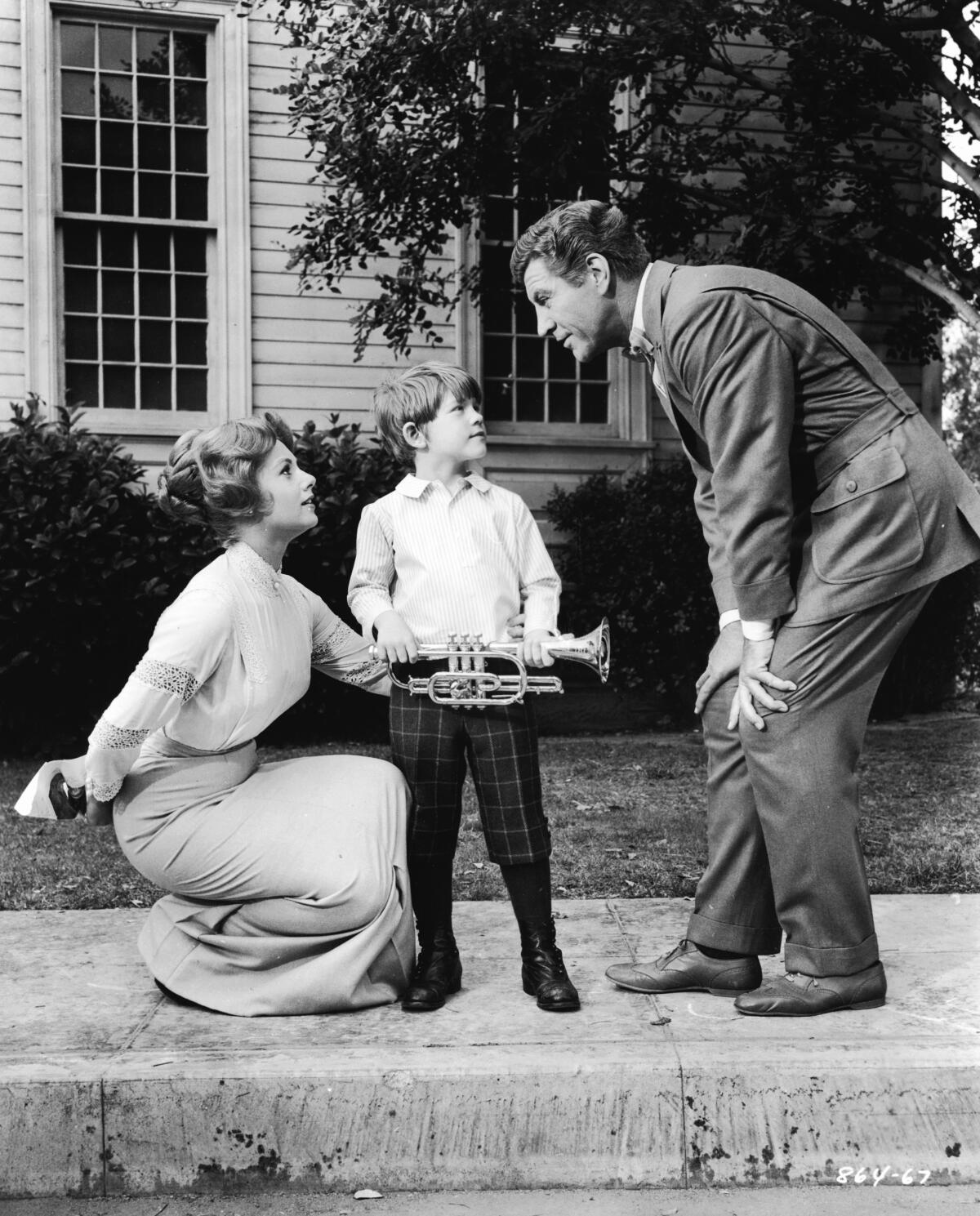A little boy holds a trumpet while a woman crouches next to him, left, and a man, right, bends down to talk to them