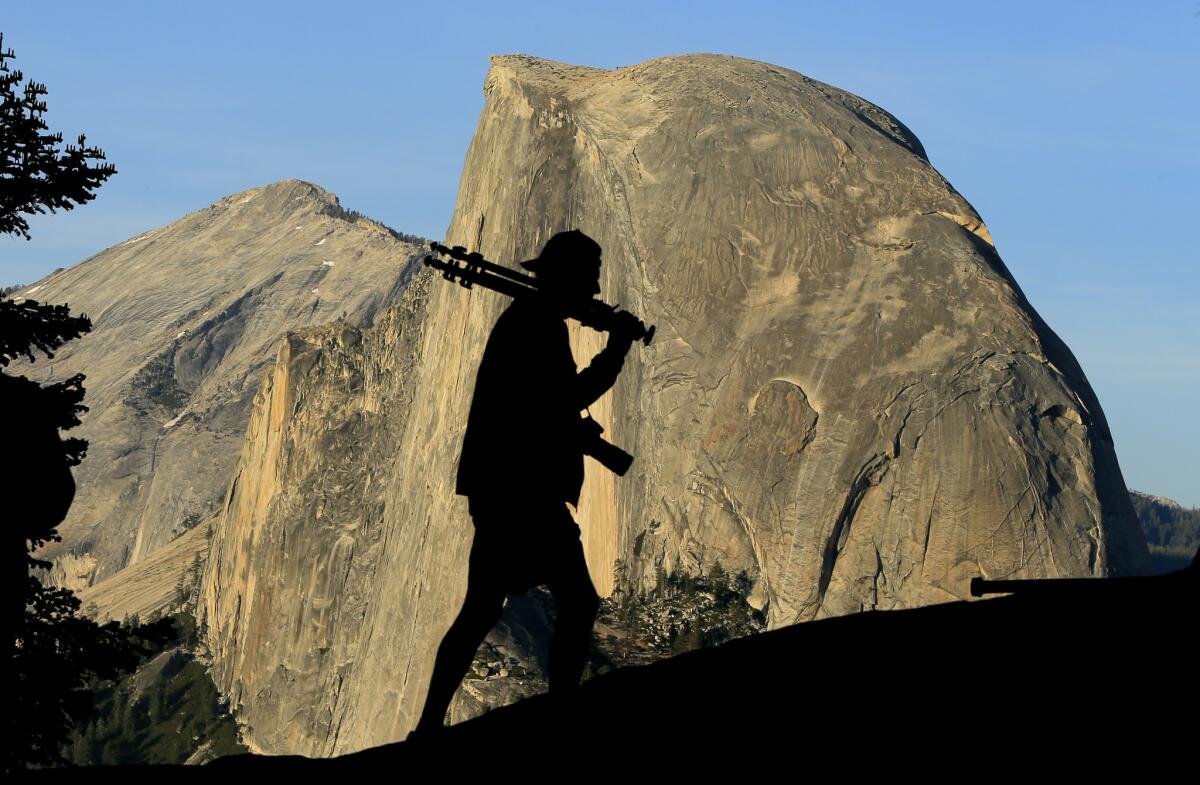 A photographer walks with his tripod over his shoulder and Half Dome in the background at Yosemite National Park.