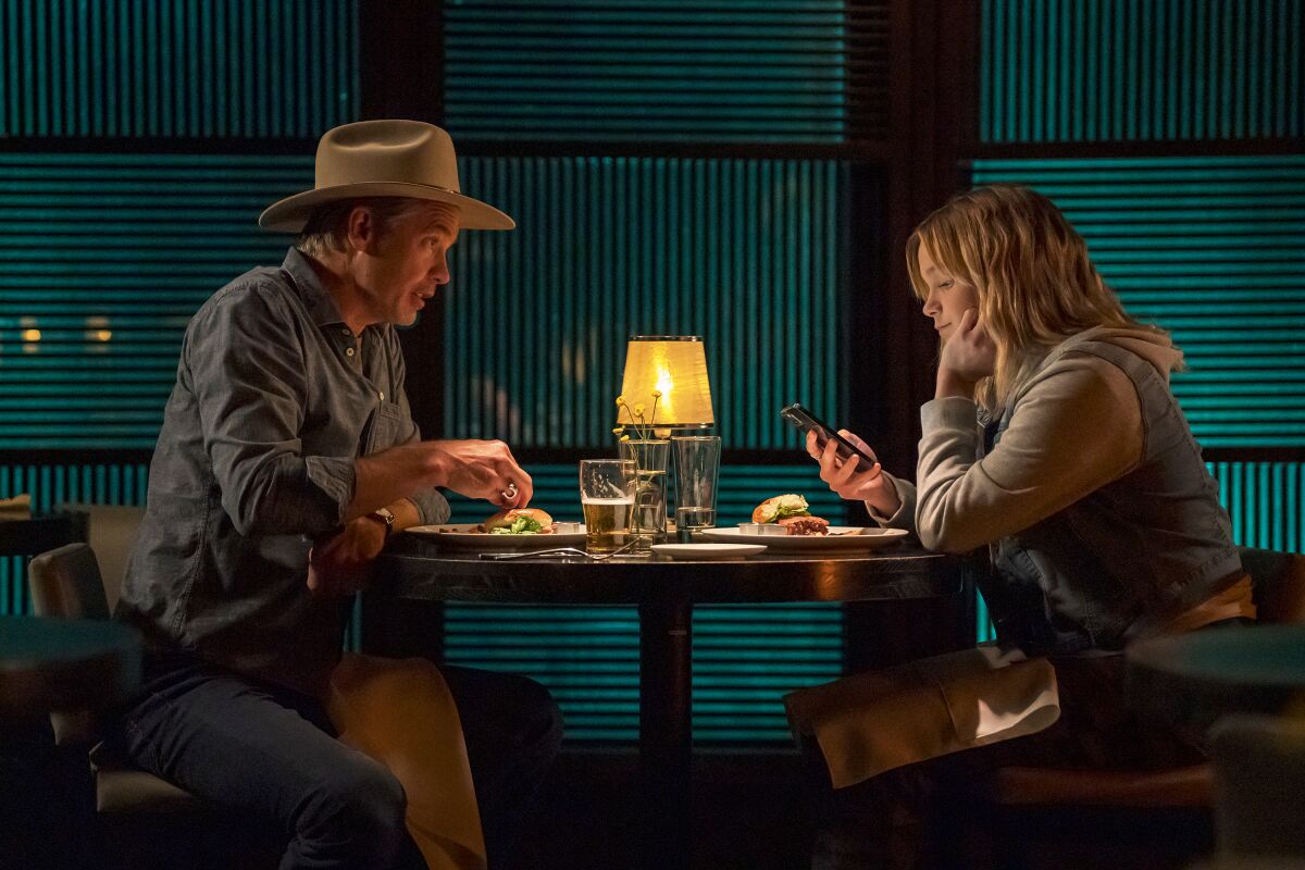 A man and his daughter sit with food on their plates in a dark dining room. She is looking at her phone.