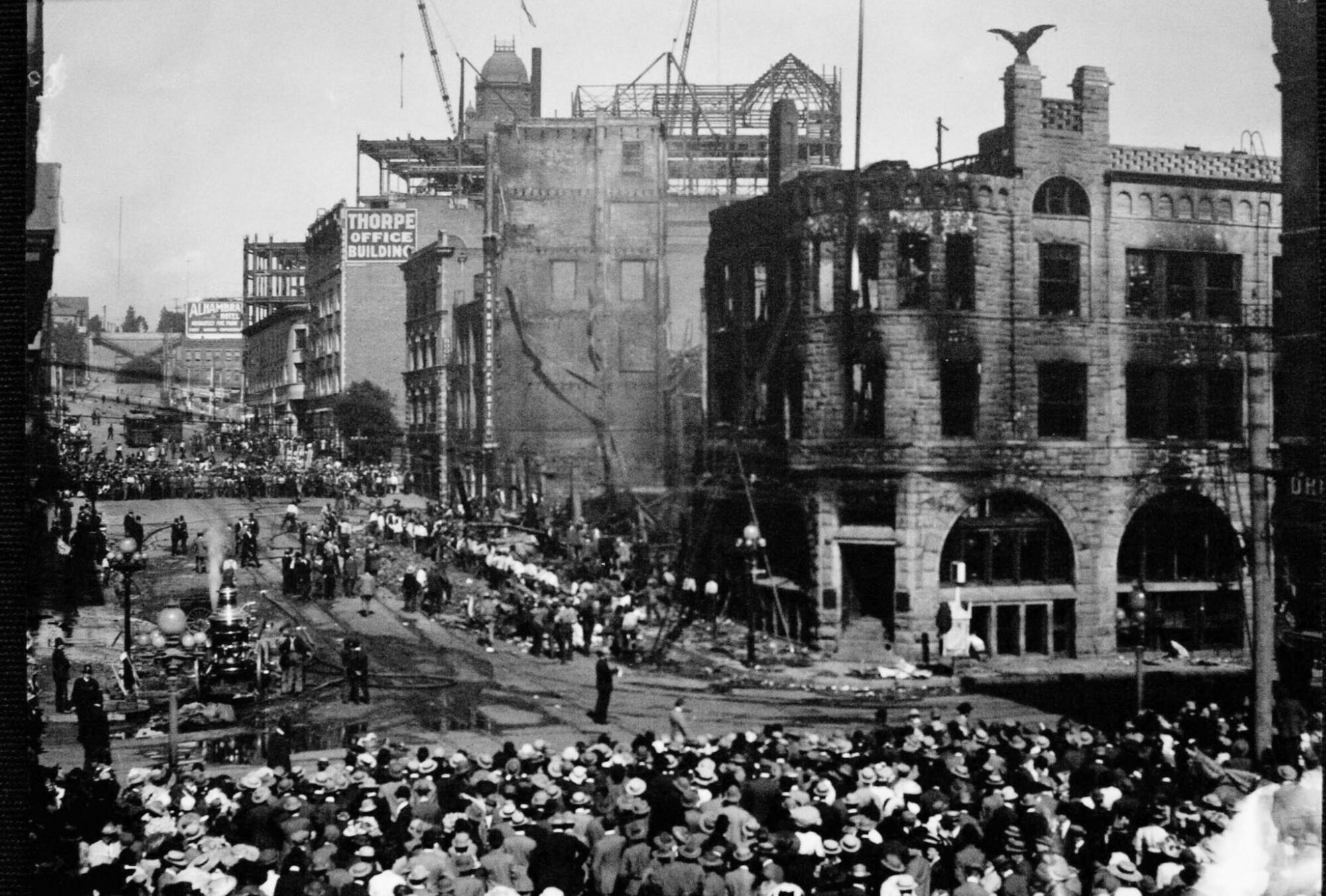 A black-and-white photo of scores of people standing across the street from a charred building with dark, blown-out windows