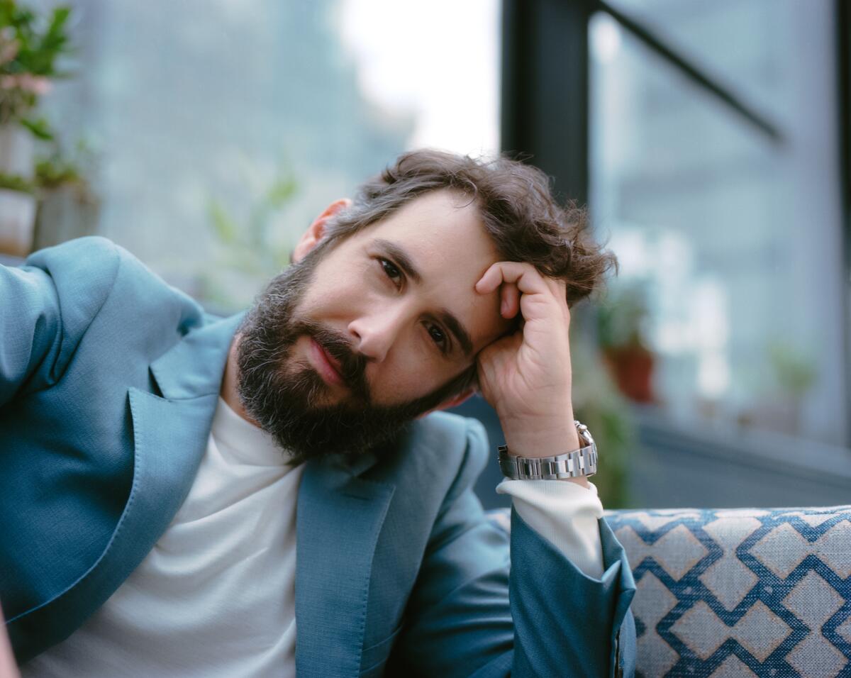 Josh Groban, in a light blue suit and wearing a beard, leans his head on his palm while sitting in a checkered banquette