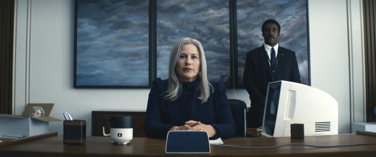 An unsmiling woman sits at a large desk with a man in a suit standing stiffly behind her in a scene from "Severance."