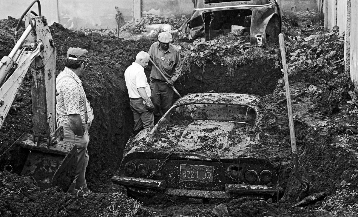 Three men stand around a sports car covered in dirt in a hole