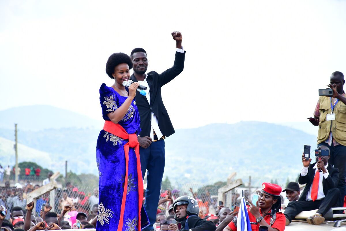 A woman in a blue dress with a red sash, and a man with his fist raised, speak to a crowd