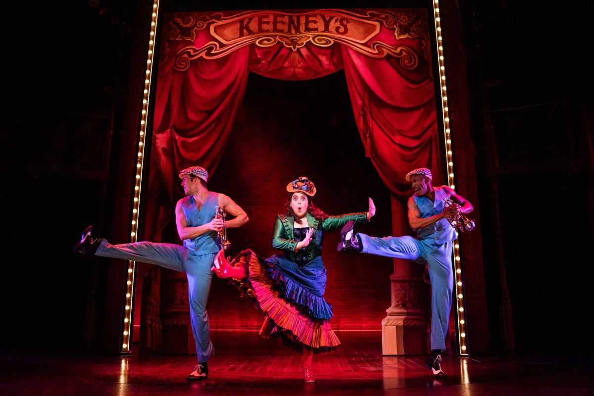 A woman and two men perform in old-timey vaudeville costumes before a red curtain.