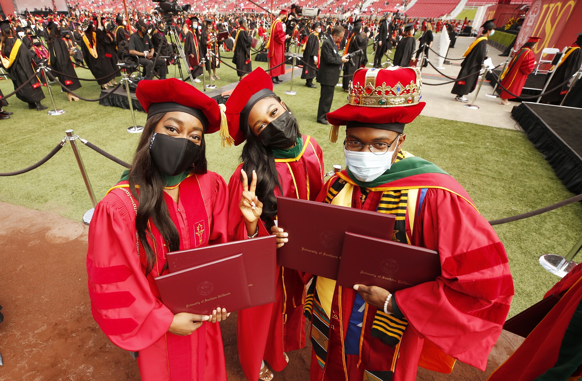 Three students in red graduation regalia hold up dual diplomas