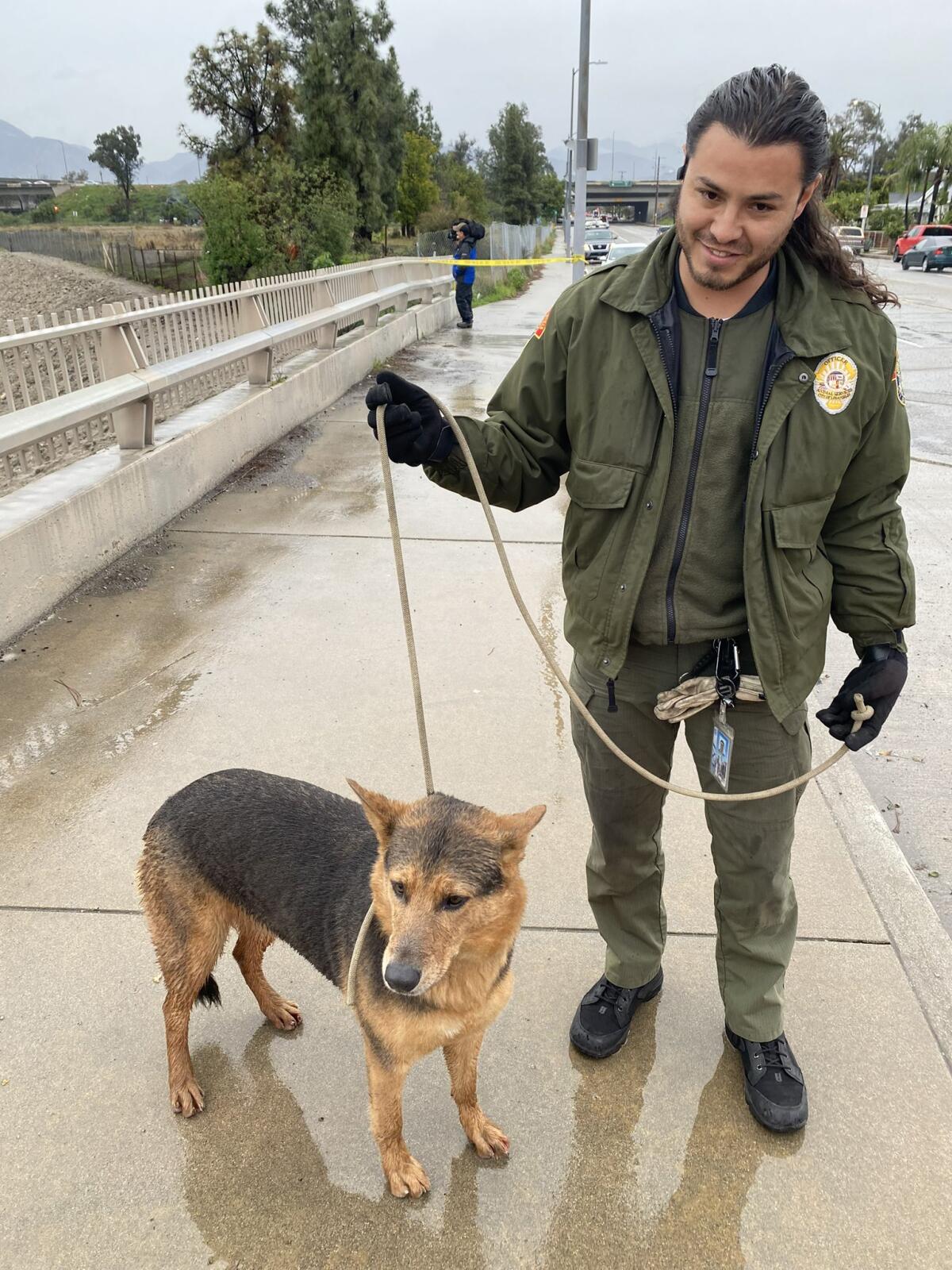 A man in a green jacket holds a brown and black dog on a leash on a rain-slicked sidewalk.