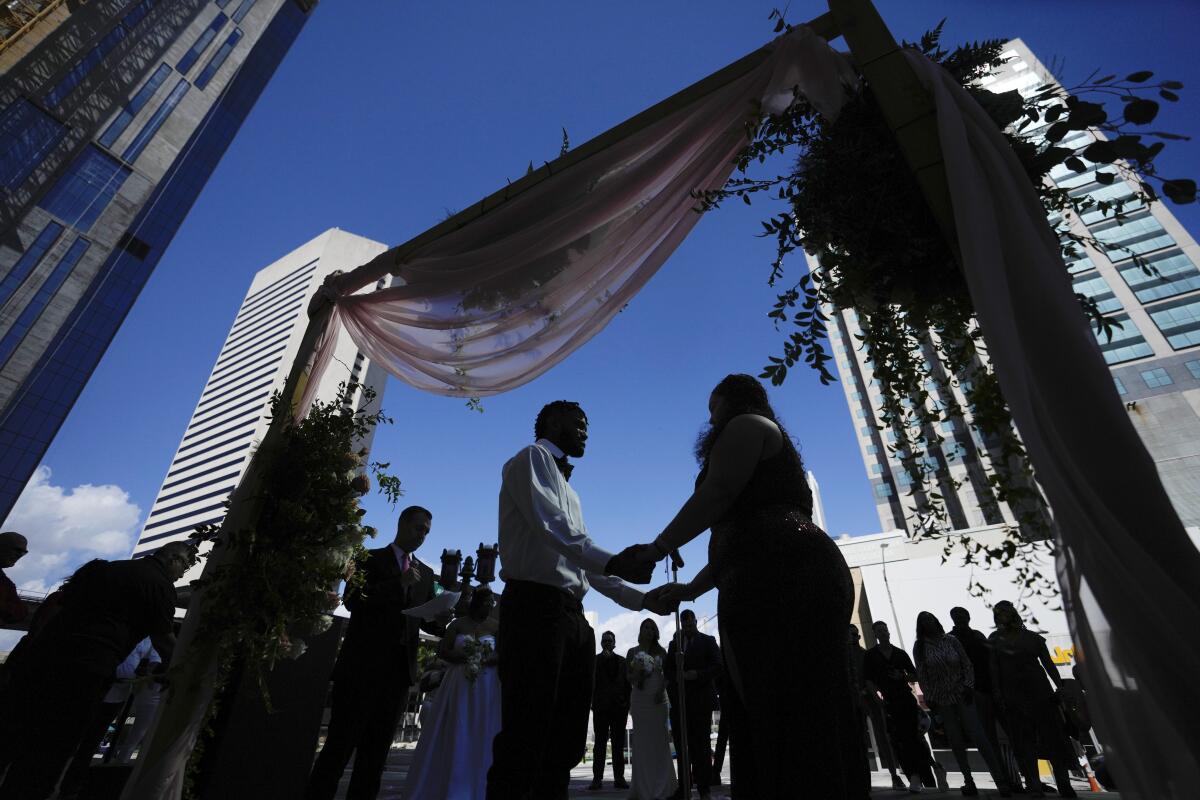 Two people hold hands under blue sky and an arch as a small crowd stands and watches against a backdrop of city skyscrapers.