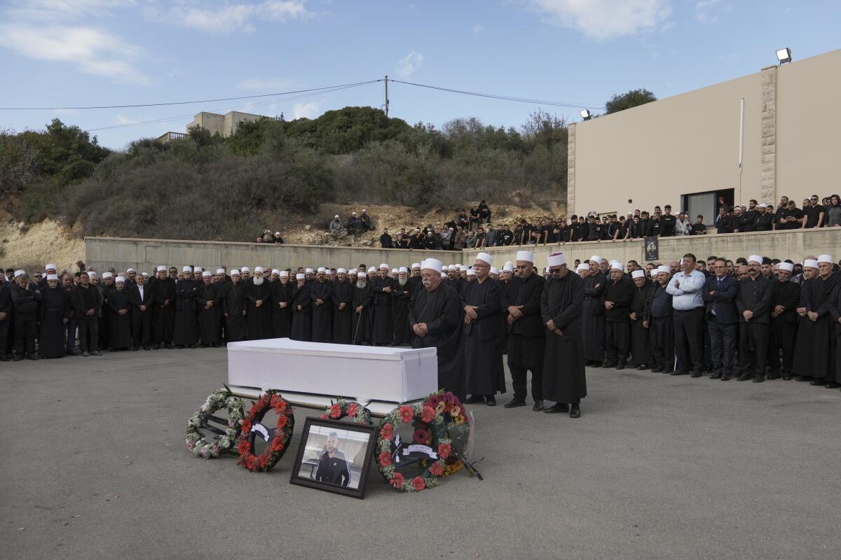Several people dressed in black with white head coverings stand near a white coffin with wreaths at its base outdoors.