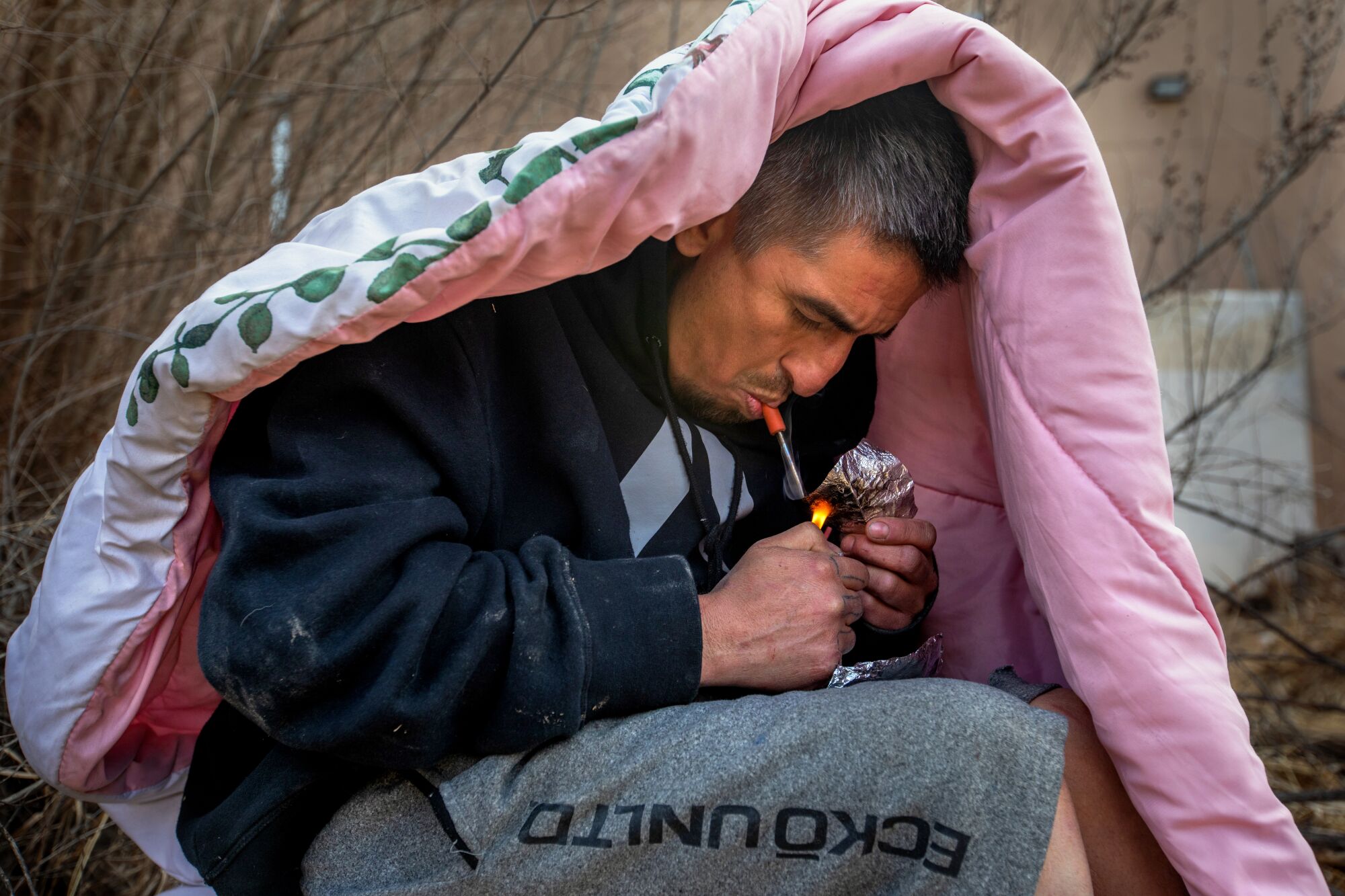 A man, sitting beneath a blanket to block the wind, smokes fentanyl.