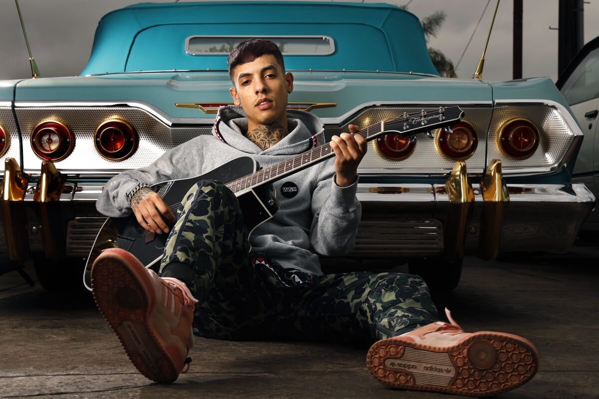 A musician sits holding a guitar behind the rear of a car.