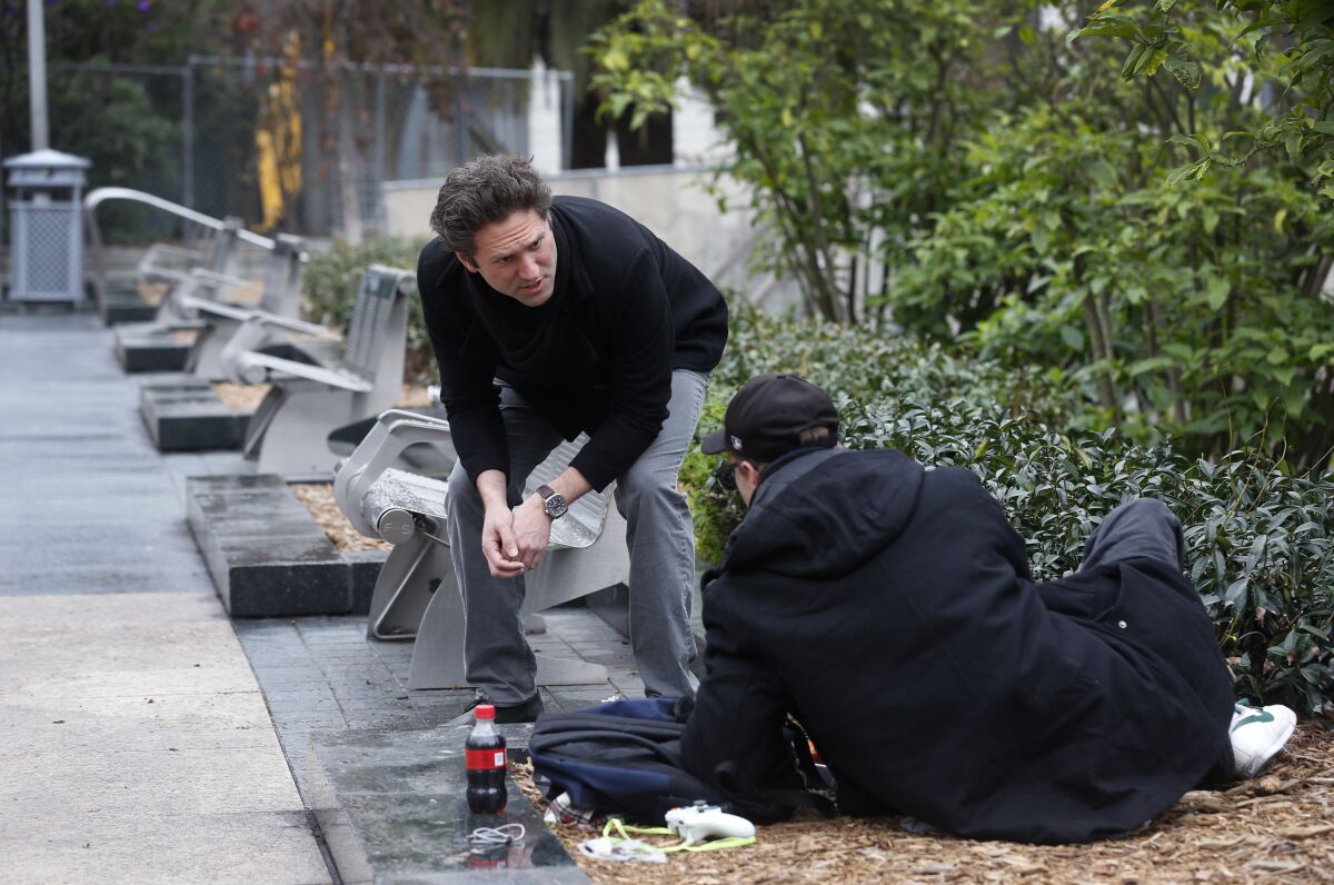 A man bends over while standing and talks with a homeless man sitting outdoors near benches.