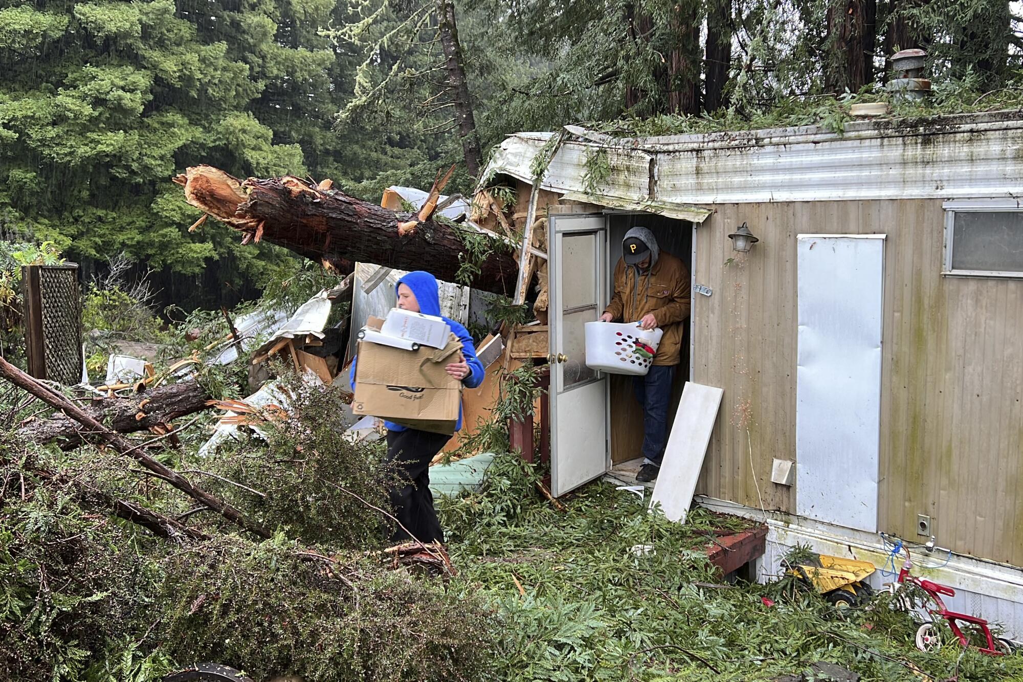 Two people walk out of a trailer surrounded by debris and tall, thick pine-tree forest.