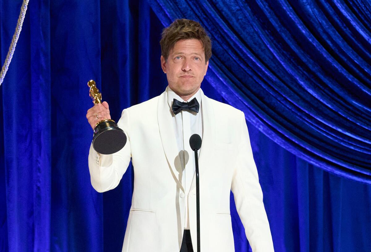 A man in a white suit and black bowtie lifts an Oscar