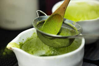 A wooden spoon pushes green powder through a sieve into a white bowl