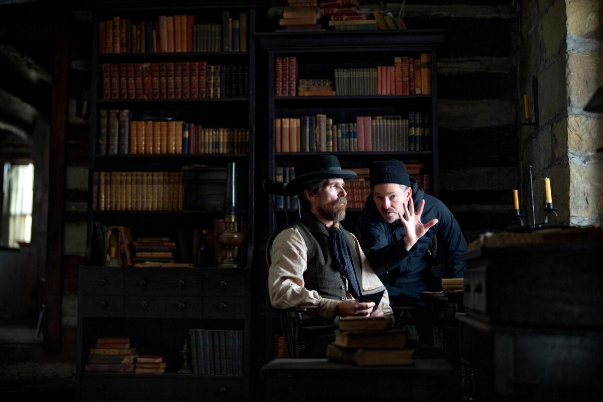 A man in Victorian-era attire talks to a director in a room filled with bookshelves.