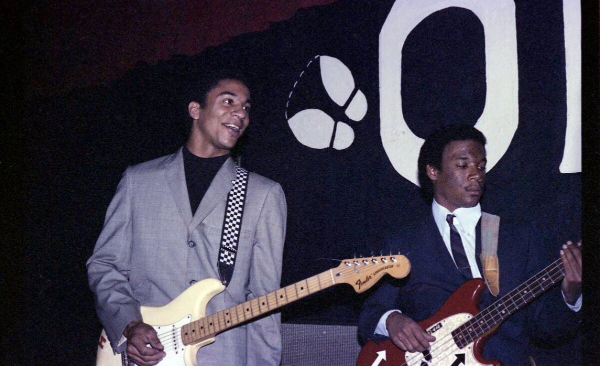 One man, left, smiles and holds a guitar while another plays the bass