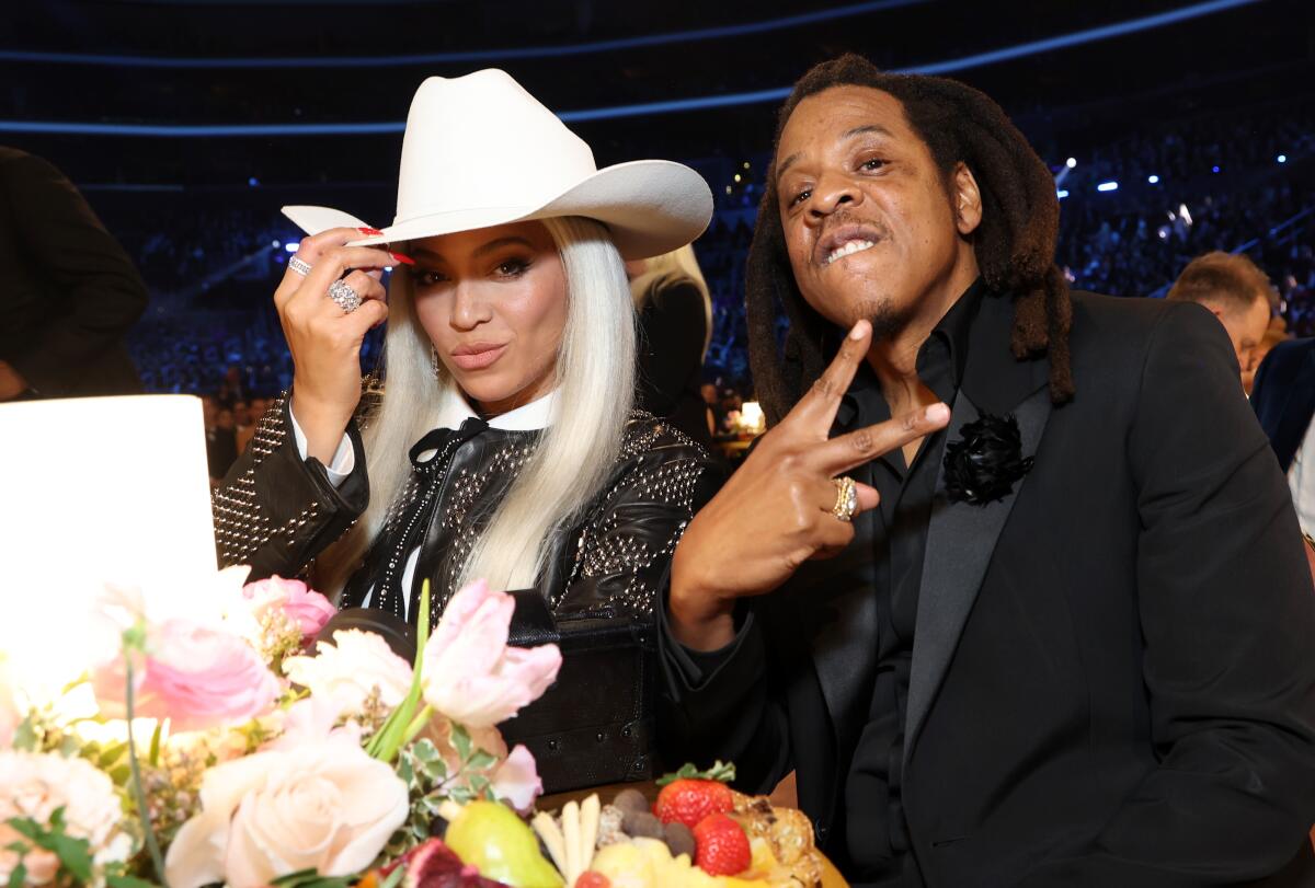 Beyoncé wearing a cowboy hat and Jay-Z wearing a black suit sit at a table with a flower arrangement on it