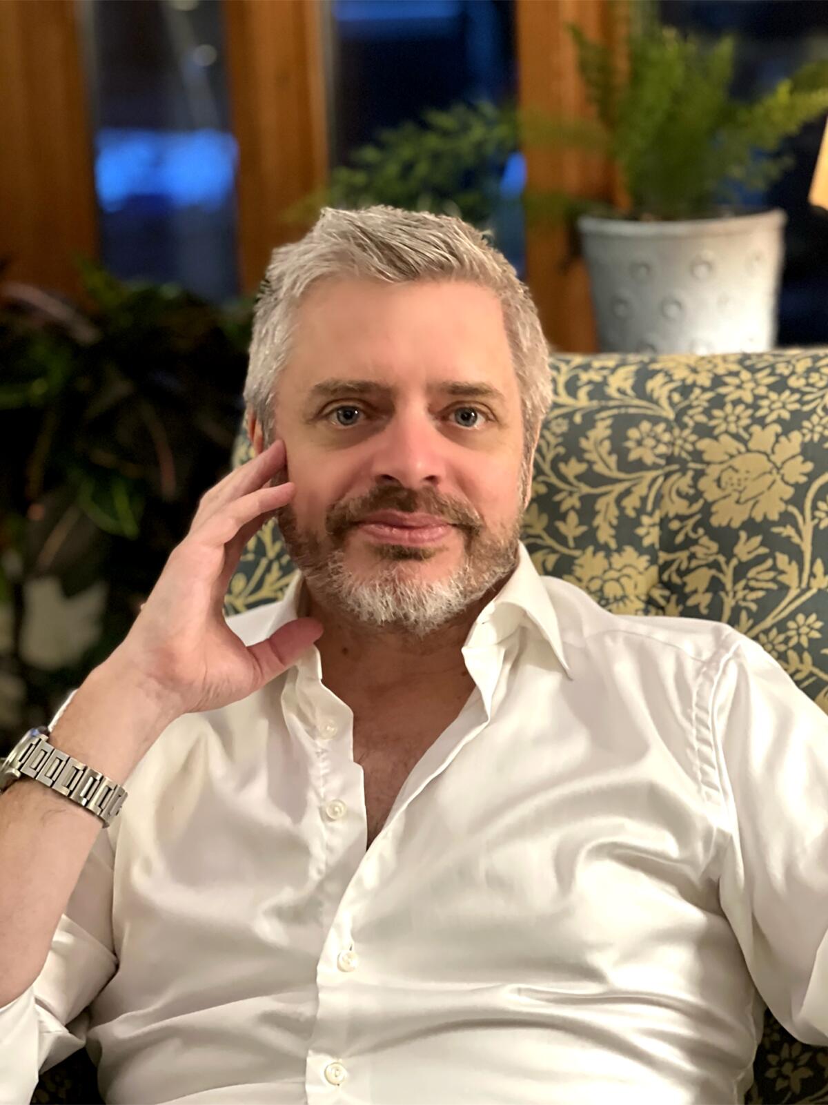 A man with a gray beard and white shirt leans his hand against his face and stares at the camera.