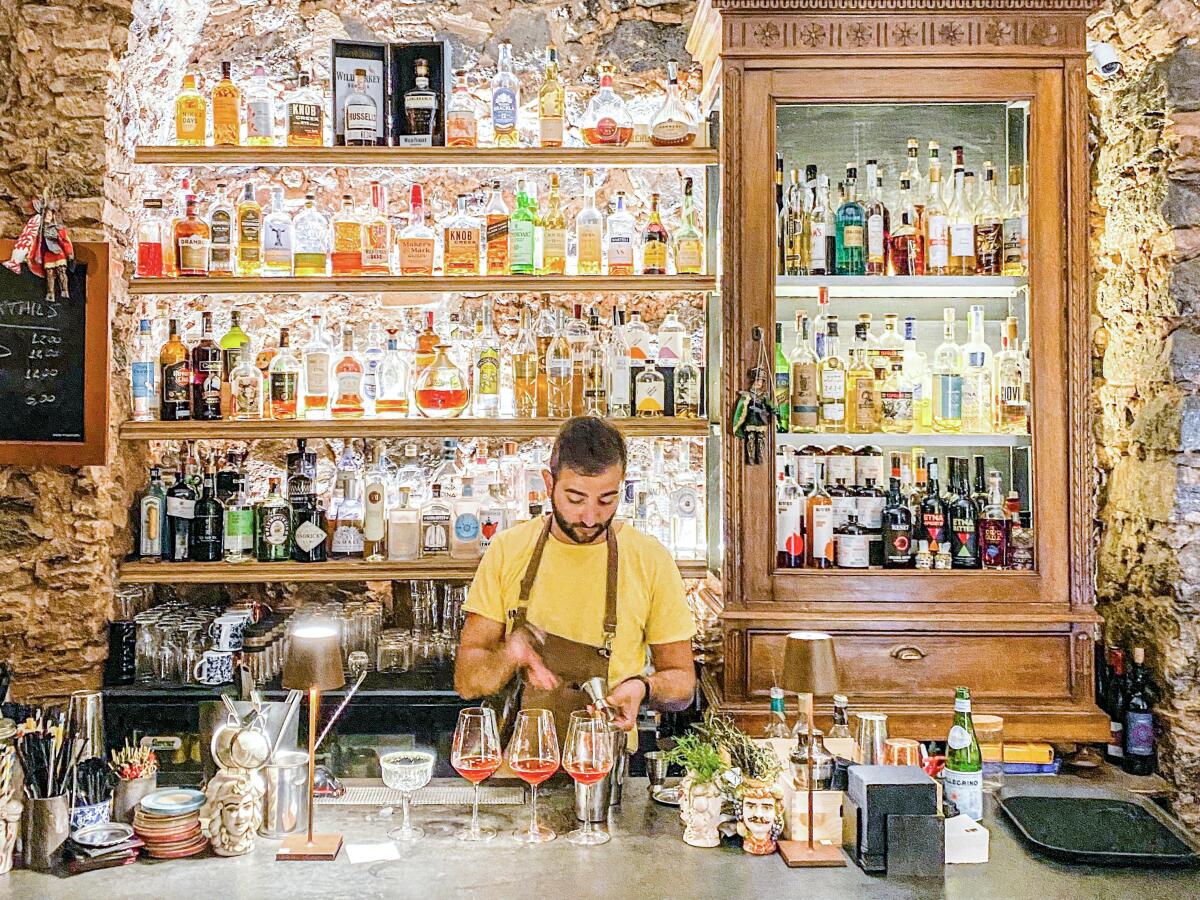 A man in an apron mixes drinks at a counter in front of shelves that hold liquor bottles. 