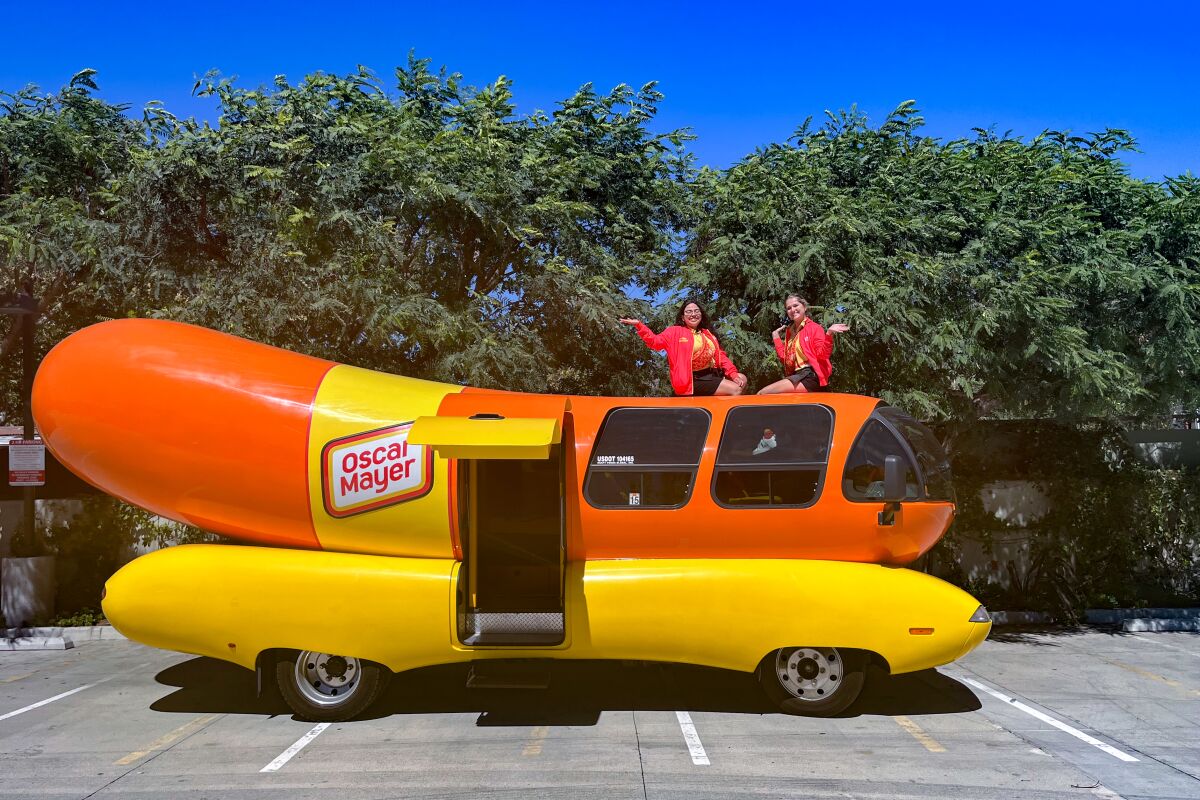 Two smiling, waving young women sit atop a yellow and red vehicle shaped like a hot dog.