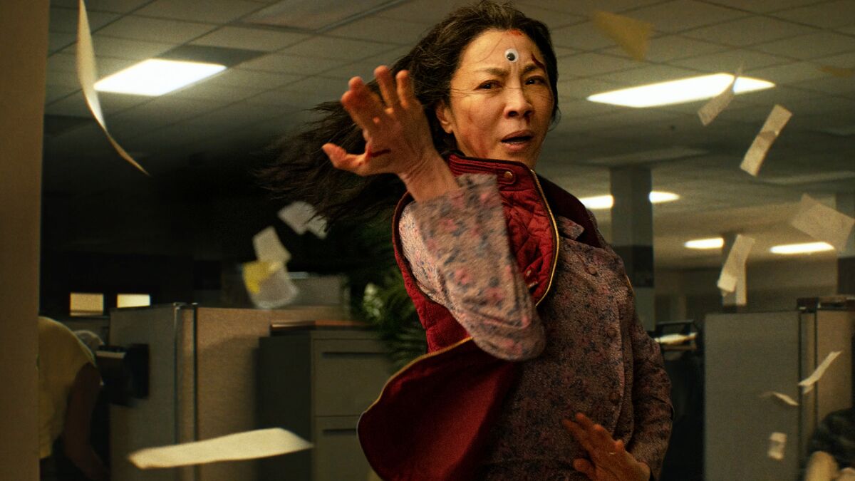 A woman with black hair and a googly eye on her forehead strikes a fighting stance in an office space.