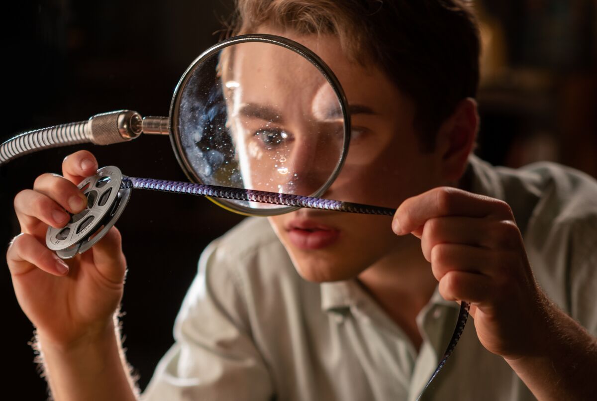 A young man examines film from a reel through a magnifying glass.