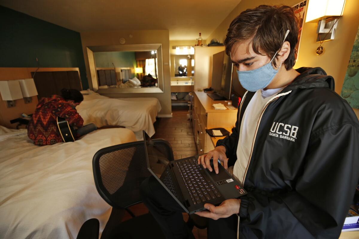 a male student holds a laptop in a hotel room as another plays guitar on a bed behind him