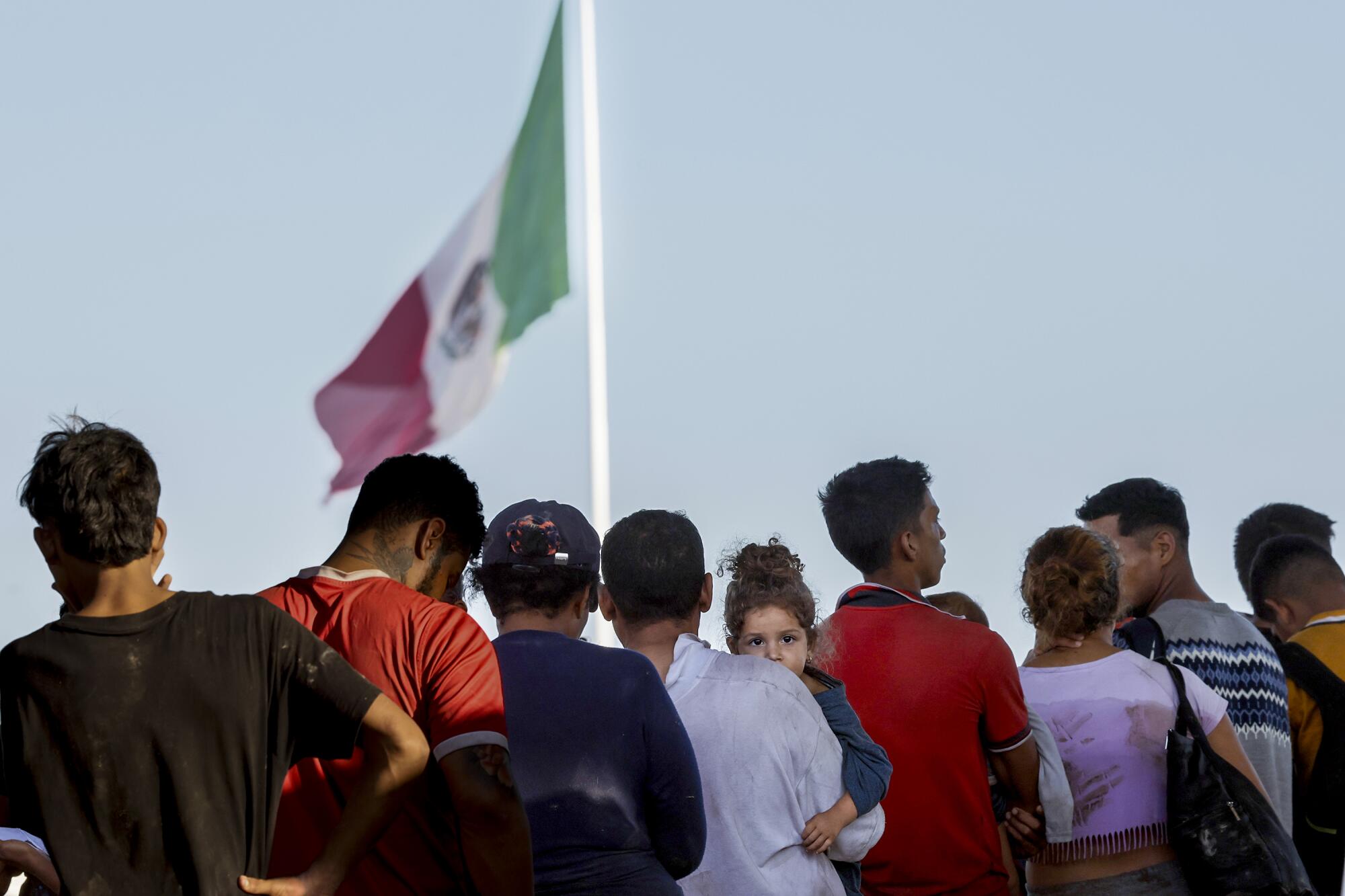 People who crossed the U.S.-Mexico border wait in a line near a flag.