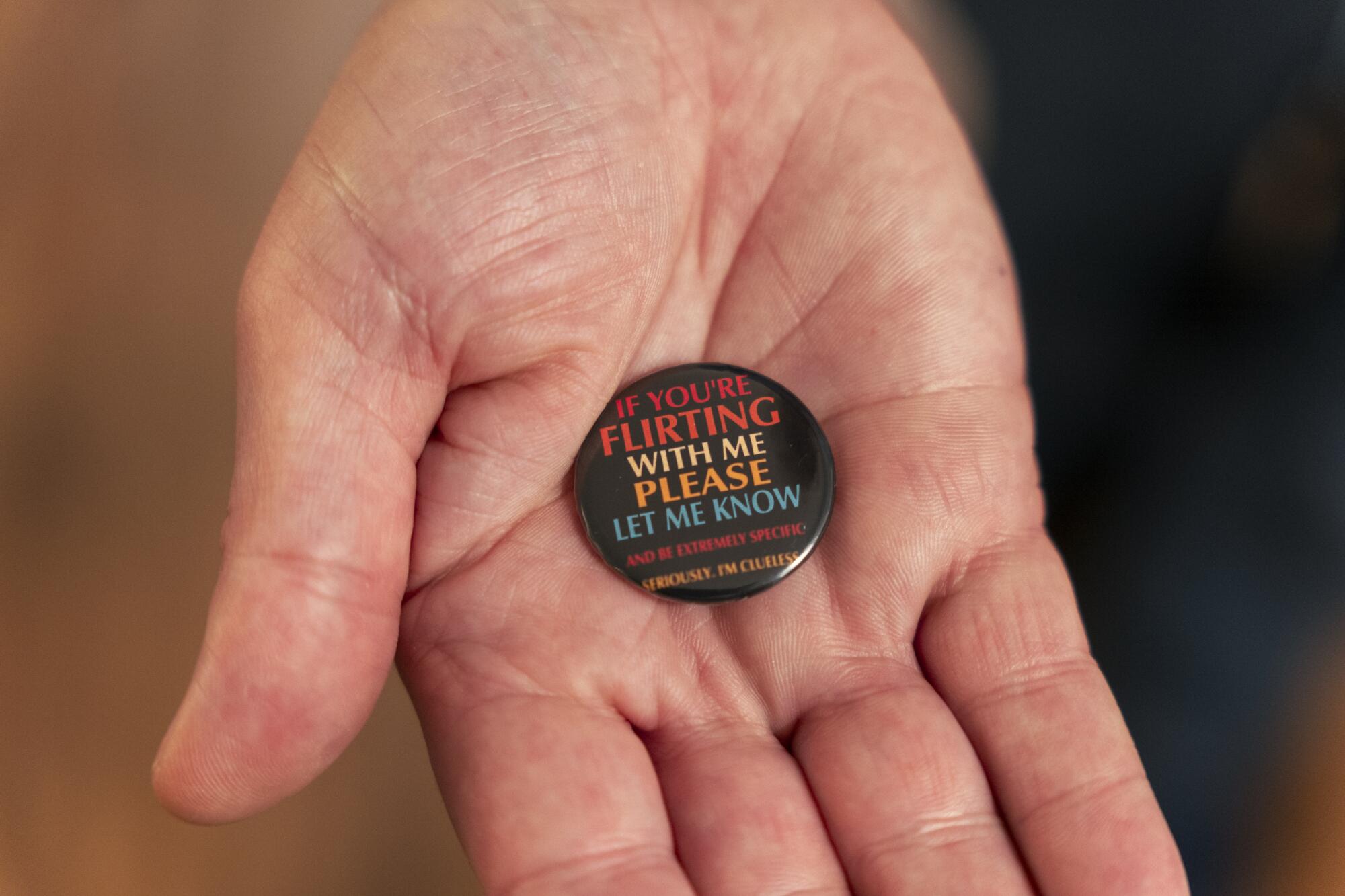 a detail of a hand holding a button that reads in part, "if you're flirting with me please let me know"