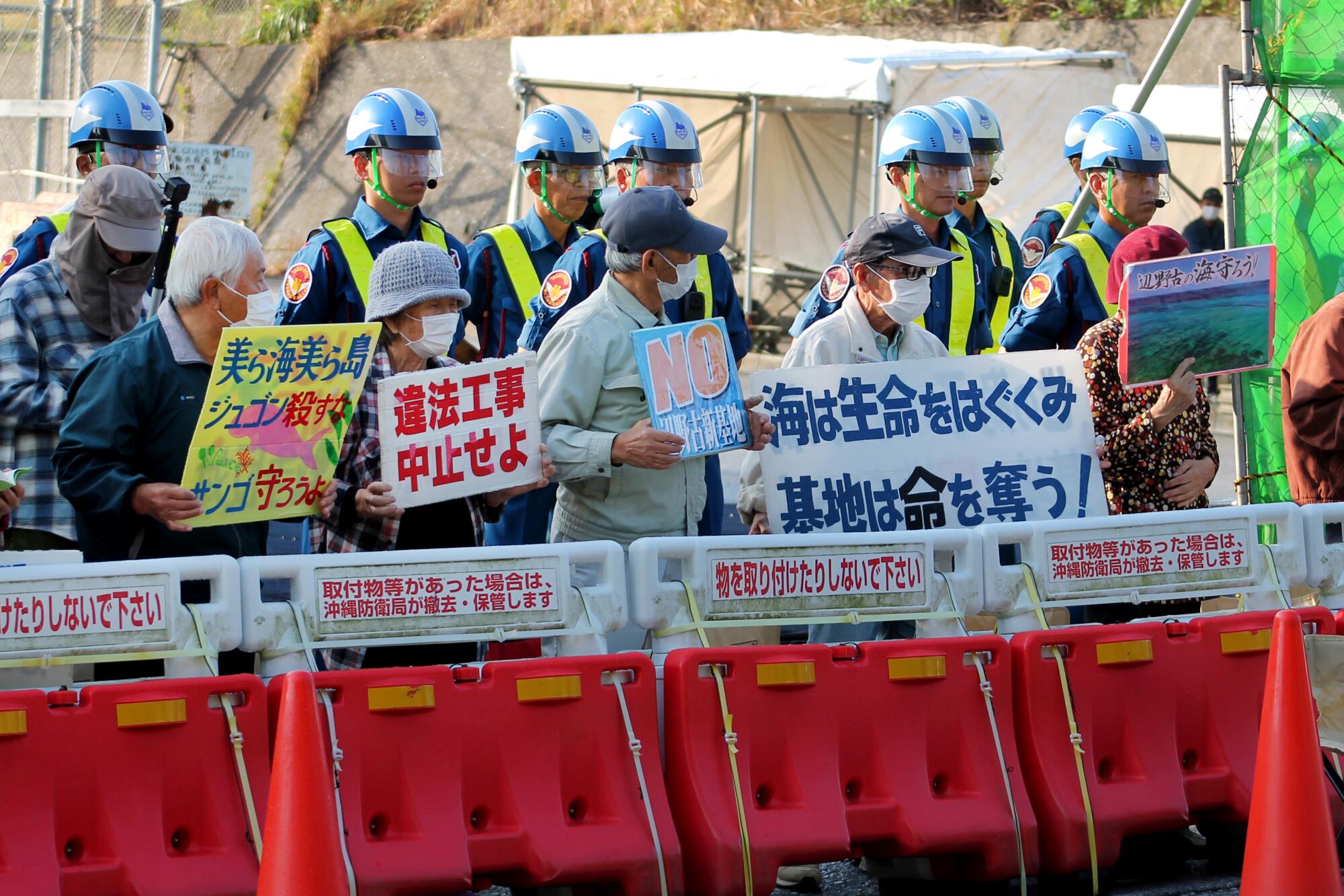 Protesters gather in front of a U.S. Marine Corps camp in Okinawa, holding signs in protest of construction and expansion.