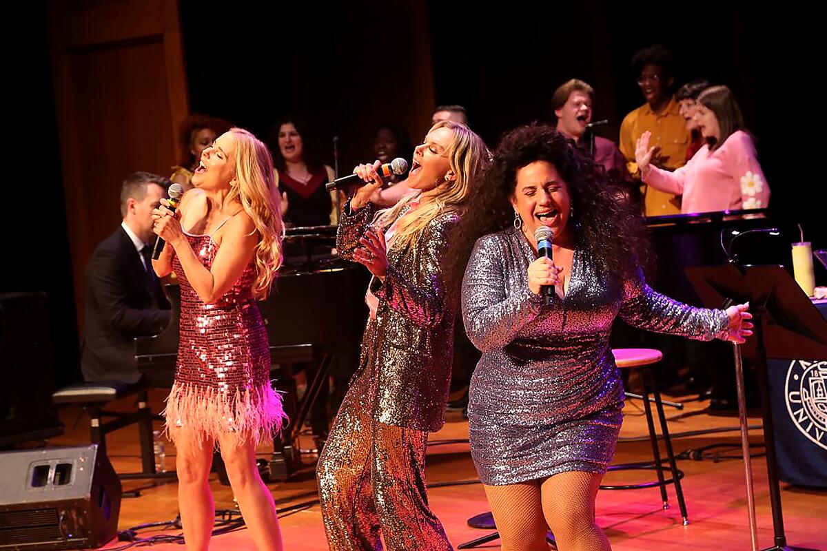 Standing on stage with hand-held mics, three women in tight, glittery short dresses belt out a song together. 