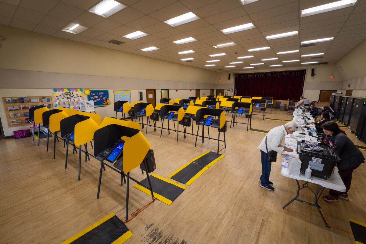 A large room with several rows of empty yellow-and-black voting booths, as workers at a long, white table help a lone voter