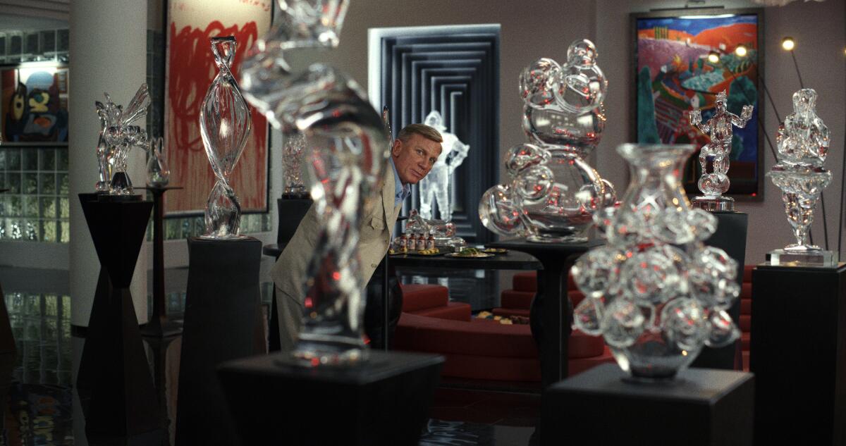 A man is seen in the background, peering at the camera past glass sculptures sitting on pedestals.