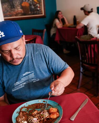 A man in a Dodgers hat eats a dish of mole at a red table in a restaurant.