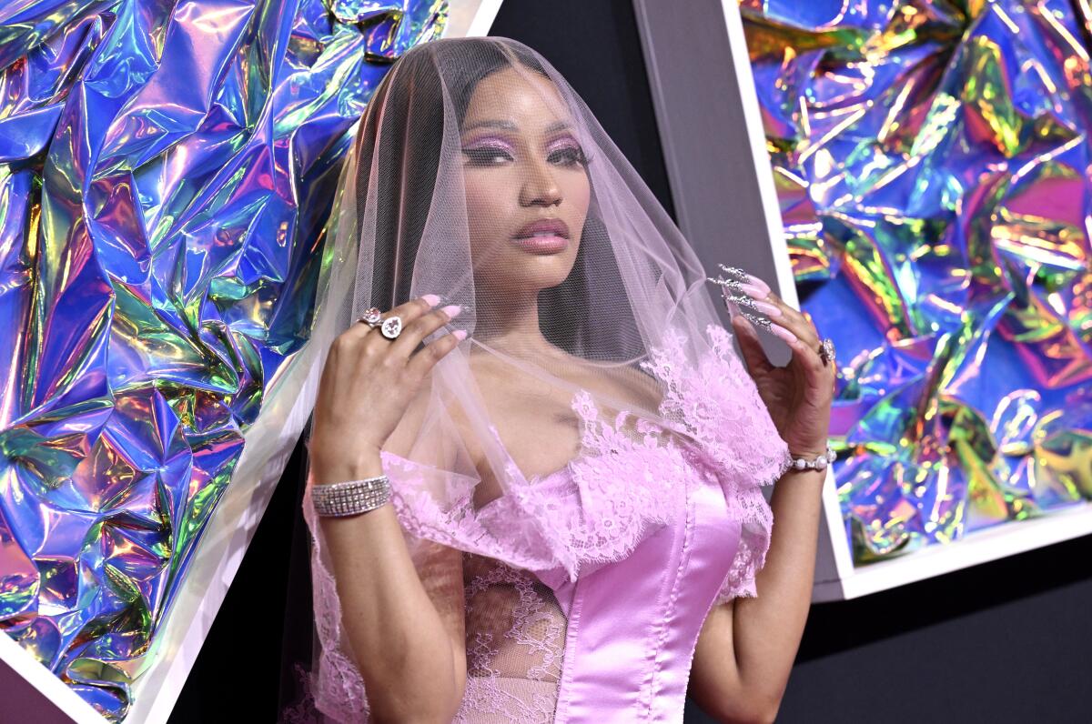 Nicki Minaj holding her hands up to her shoulders wearing a pink satin corset and a pink veil