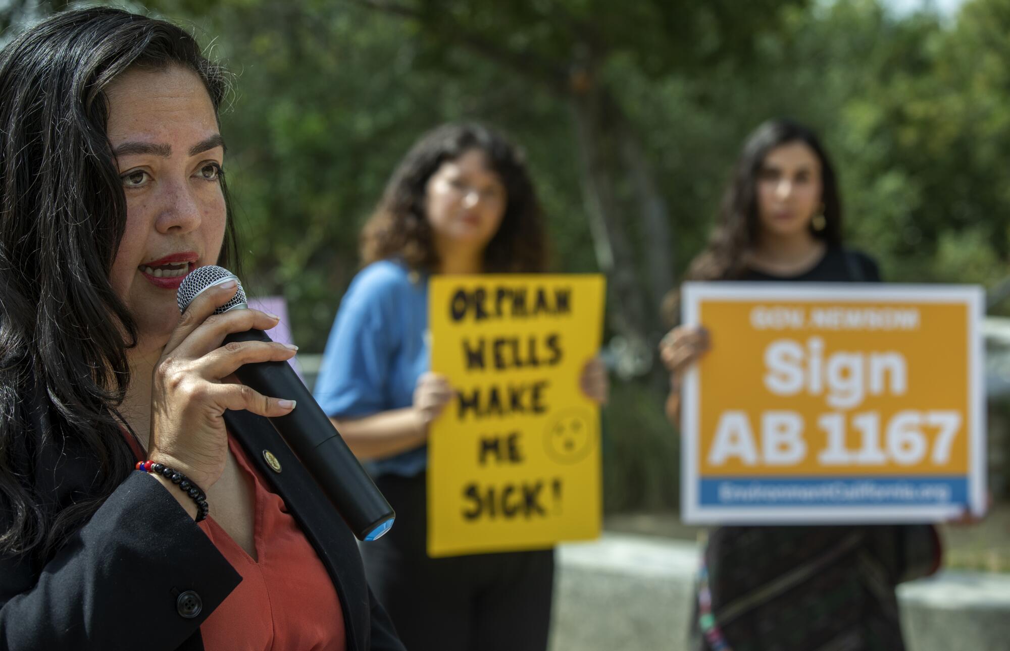 A woman speaks into a microphone as two other women hold signs.