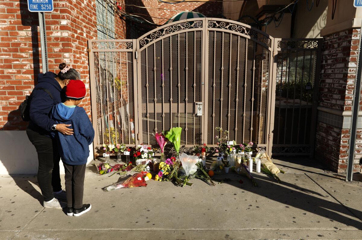A woman and child stand in front of a gate with flower arrangements and candles form a memorial.