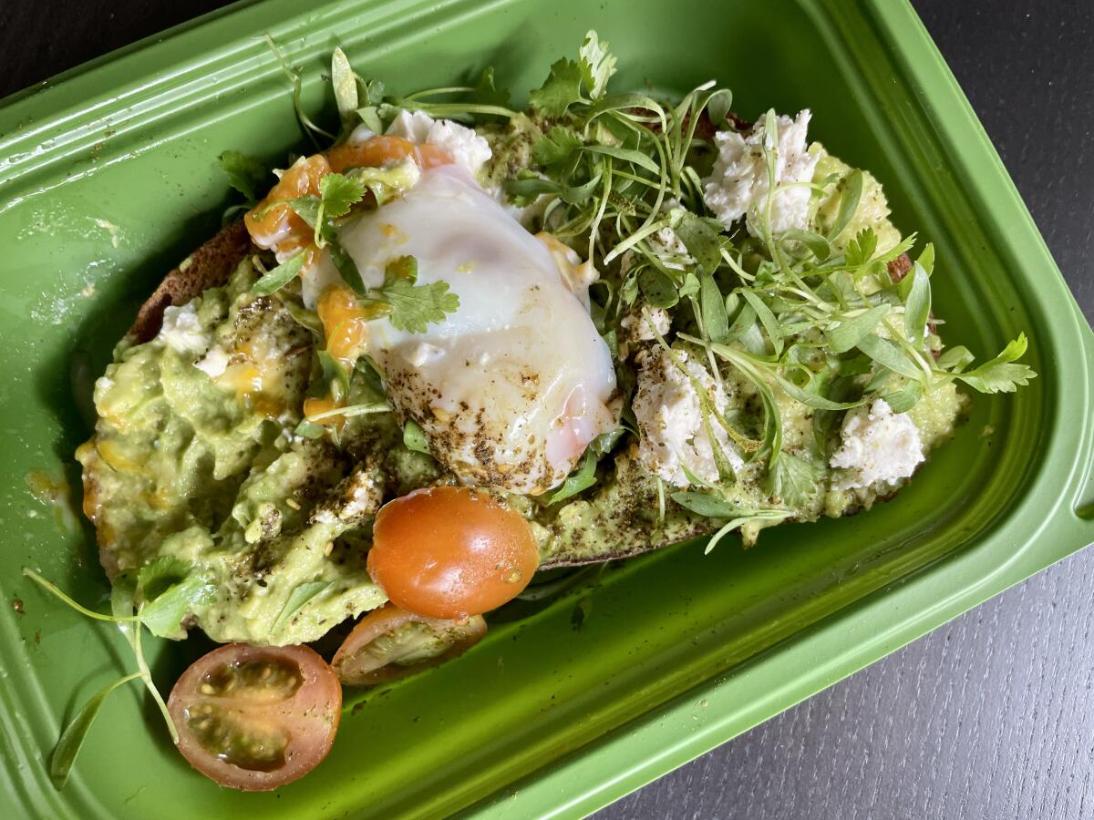A green container holds a piece of avocado toast with microgreens and cherry tomatoes