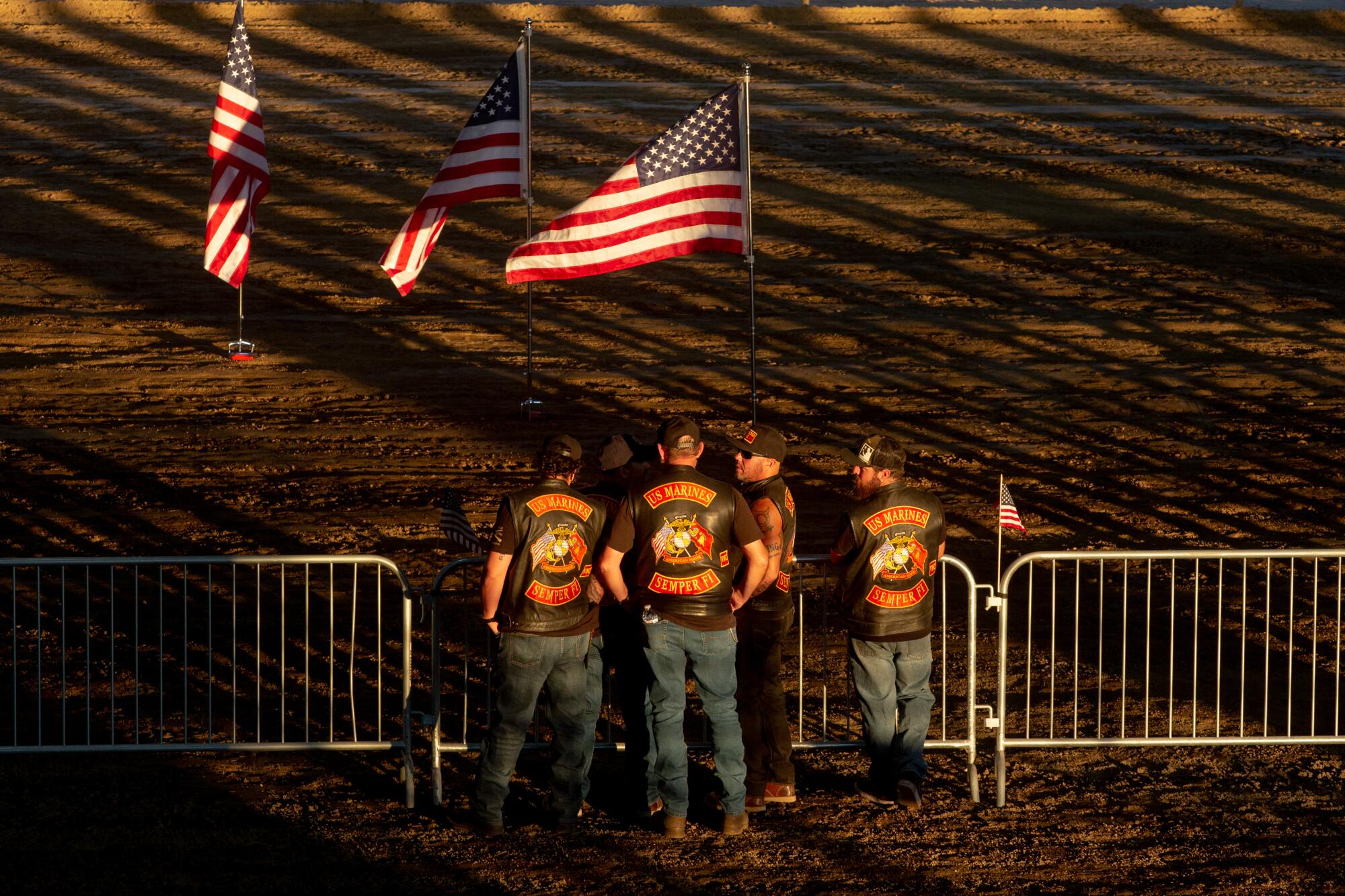 Some men in leather vests stand outside near U.S. flags as the sun sets