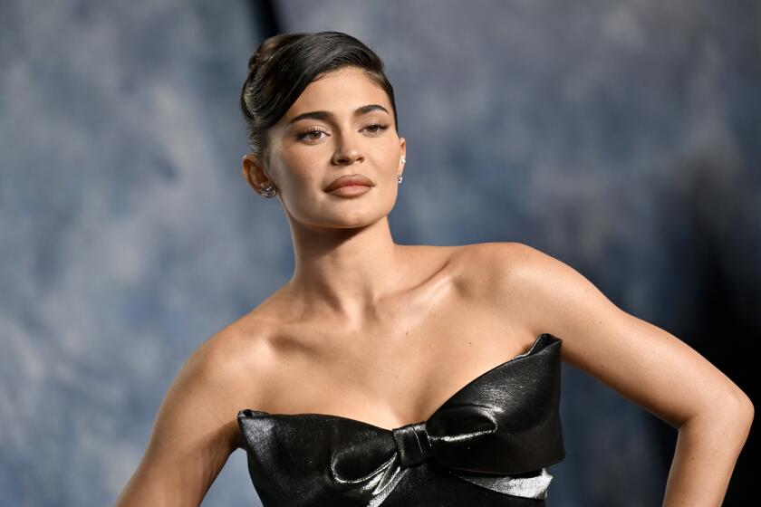 Kylie Jenner in an updo and a shiny black, strapless dress posing for pictures in front of a grey background